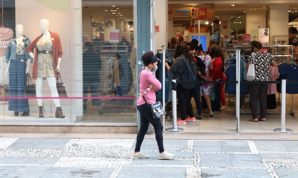 Mulher caminha de máscara pela calçada em frente a uma loja aberta e com pessoas dentro do estabelecimento.