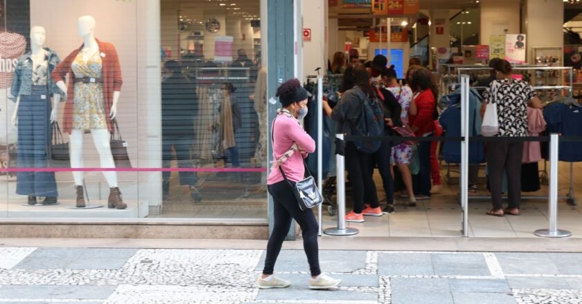 Mulher caminha de máscara pela calçada em frente a uma loja aberta e com pessoas dentro do estabelecimento.