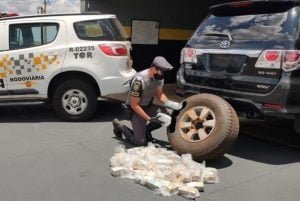 Com luvas, policial militar retira pacotes de dinheiro de dentro do pneu ao lado de uma viatura do carro que fazia o transporte