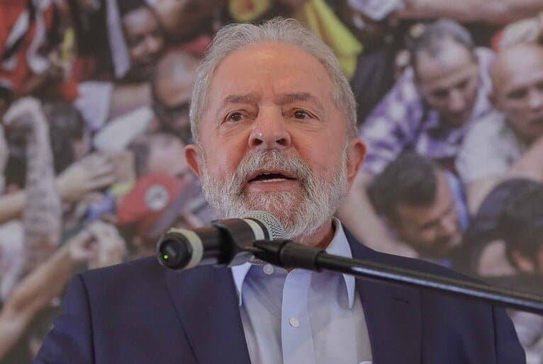 Lula fala ao microfone. Atrás de lula, um painel exibe imagem de pessoas aglomeradas.