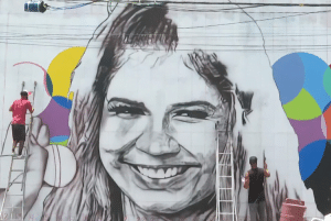 Marília Mendonça sorrindo e artista trabalhando a pintura no mural de 23 metros