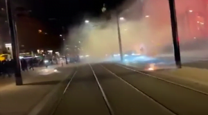 Bombas explodem no meio na rua. Fumaça de fogos se espalham pela rua.