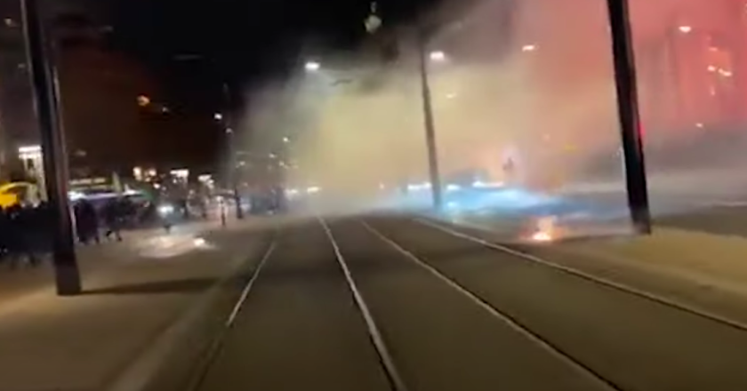 Bombas explodem no meio na rua. Fumaça de fogos se espalham pela rua.