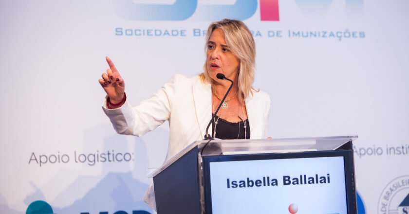 Isabella Ballalai fala ao microfone. É uma mulher branca, loira, cabelos perto dos ombros. Veste um blazer branco, com blusa preta por baixo e está com um óculos pendurado no pescoço. Com a mão direita, aponta para o alto. Ao fundo, um banner com a logomarca da Sociedade Brasileira de Imunizações.