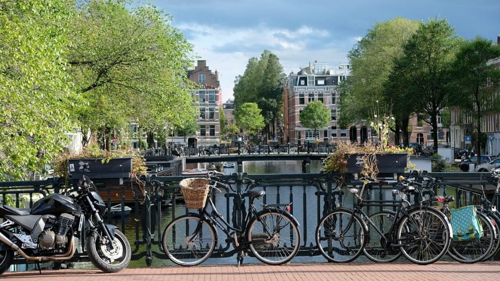 Canal em Amsterdam, na Holanda, visto de cima de uma ponte. Foto mostra bicicletas estacionadas sobre a ponte. Ao fundo, árvores com a copa verde e alguns prédios.