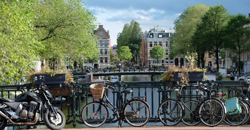 Canal em Amsterdam, na Holanda, visto de cima de uma ponte. Foto mostra bicicletas estacionadas sobre a ponte. Ao fundo, árvores com a copa verde e alguns prédios.