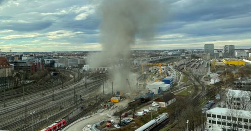 Imagem panorâmica da região onde ocorreu a explosão, perto de uma linha de trem. Trem aparece parado. Fumaça subindo. Céu encoberto por nuvens. Região é cercada por muitos prédios.