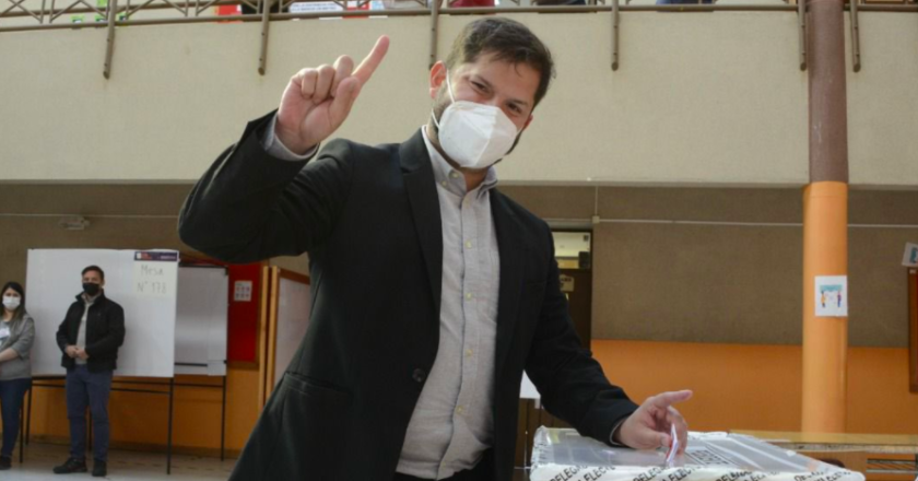 Gabriel Boric, homem de pele branca, usa blazer preto e camisa social clara. Com máscara no rosto, deposita foto em urna de votação e com a outra mão aponta o dedo indicador para o alto.