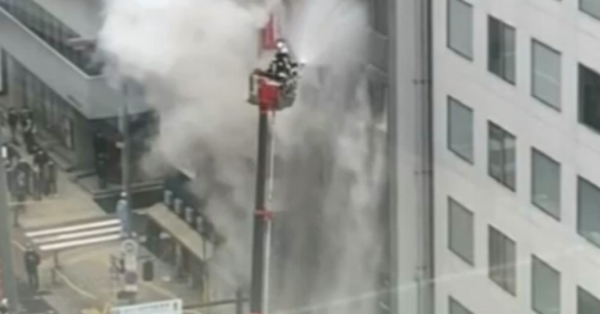 Escada de combate a incêndio dos bombeiros, com homem do corpo de bombeiros no alto, lança água no local do incêndio. Ao fundo, muita fumaça que sai do edifício.
