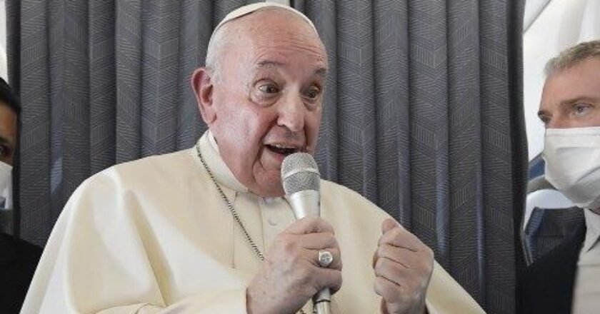 Papa Francisco segurando o microfone. Enquanto fala, é observado por dois assessores que usam máscara e terno e gravata.