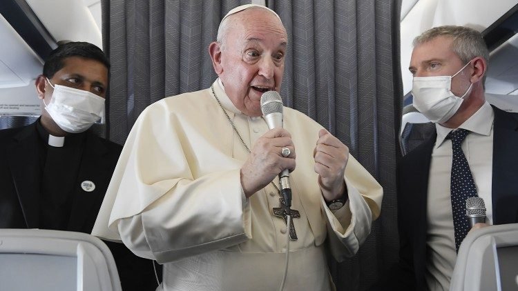 Papa Francisco segurando o microfone. Enquanto fala, é observado por dois assessores que usam máscara e terno e gravata.