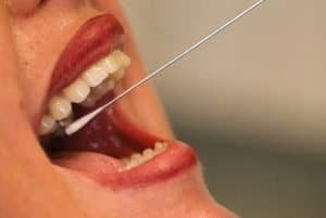 Foto em close na boca aberta de uma pessoa e haste de coleta de pcr dentro da boca.