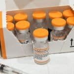 Frascos da vacina da pfizer com tampas na cor laranja. No canto esquerdo parte de um seringa.
