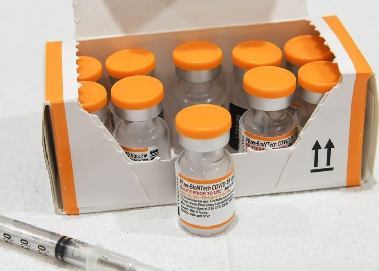 Frascos da vacina da pfizer com tampas na cor laranja. No canto esquerdo parte de um seringa.