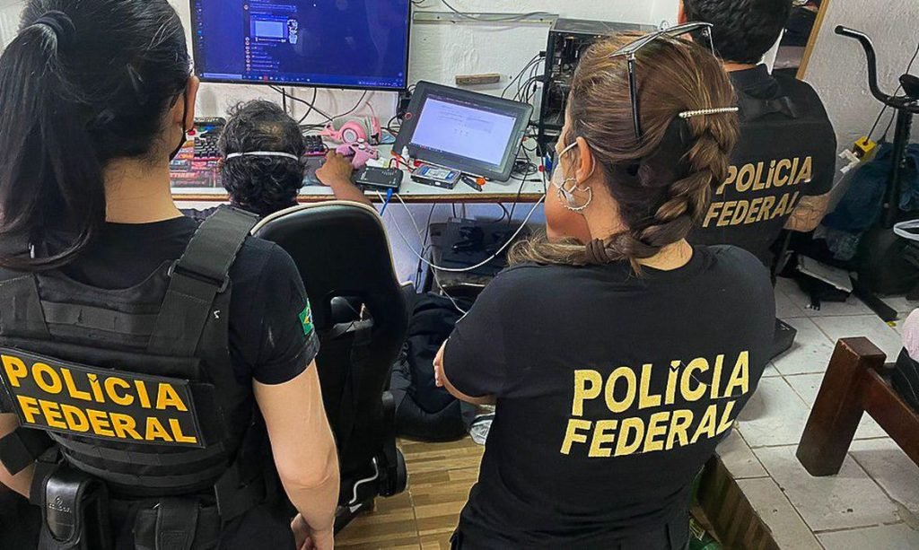Agentes da Polícia Federal observam computador. Na camisa dos policiais há o nome da Polícia Federal.
