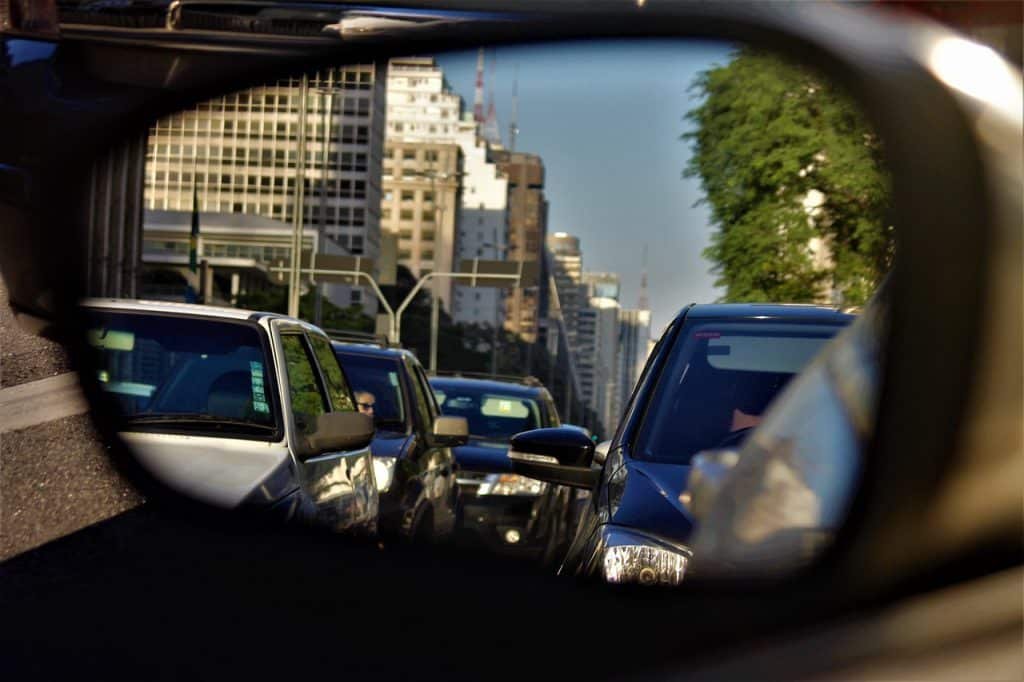 Espelho retrovisor de carro reflete veículos enfileirados em via de São paulo. Ao fundo, topo de prédios sem definição.
