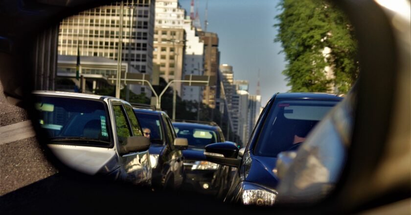 Espelho retrovisor de carro reflete veículos enfileirados em via de São paulo. Ao fundo, topo de prédios sem definição.