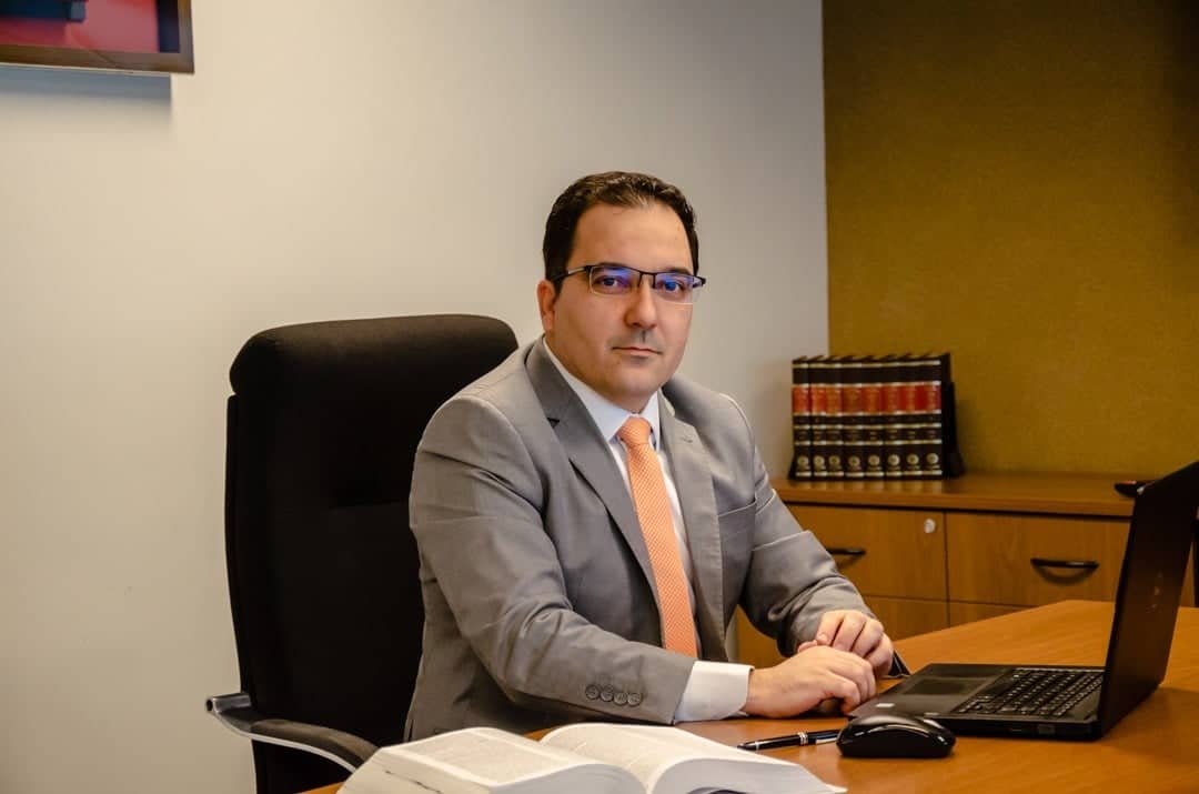 Angelo Ambrizzi, homem de pele branca, cabelos curtos, com óculos de grau. Está sentado à mesa do escritório, com computador e livro abertos à sua frente. Veste terno e gravata.