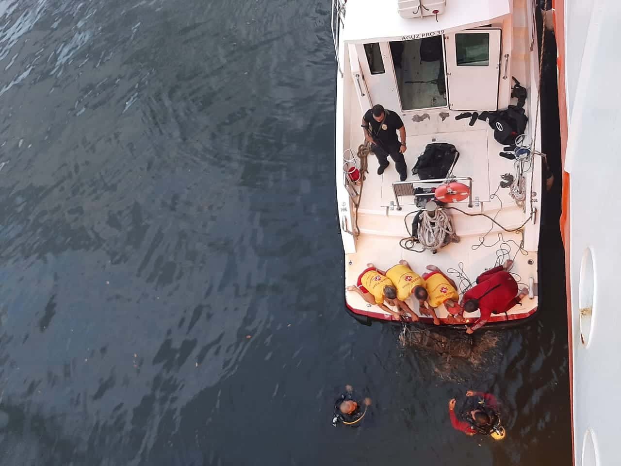 Foto vista de cima, mostra mergulhadores e agentes dentro de um barco enquanto outros mergulhadores que estão dentro  d'àgua.