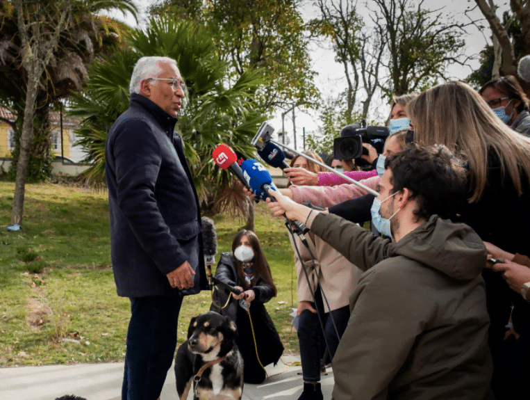 António Costa, homem branco e cabelos brancos, com óculos de grau, fala a jornalistas que aparecem no canto direito da foto segurando microfones. Dois cachorros acompanham o momento perto de António.