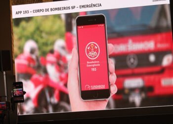 Telão, durante entrevista coletiva, mostra tela de celular com o aplicativo que permite acionar bombeiros e pm. Ao fundo da imagem há um caminhão dos bombeiros e motos da corporação.