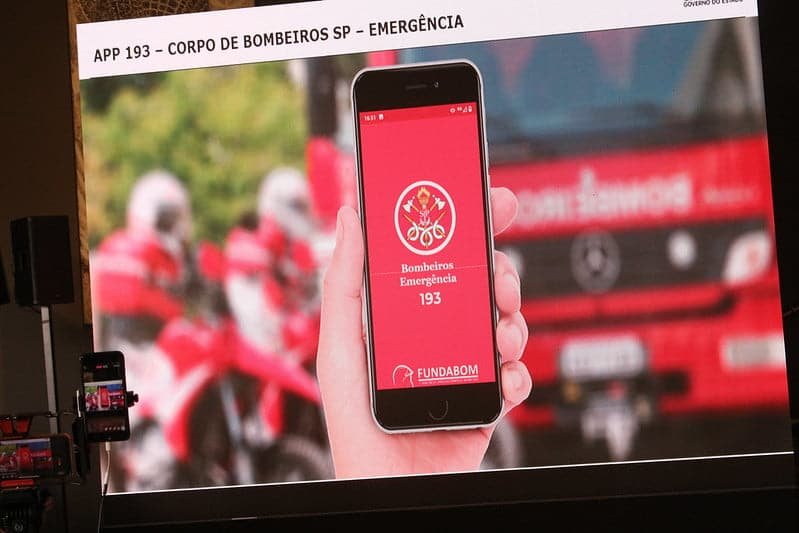 Telão, durante entrevista coletiva, mostra tela de celular com o aplicativo que permite acionar bombeiros e pm. Ao fundo da imagem há um caminhão dos bombeiros e motos da corporação.