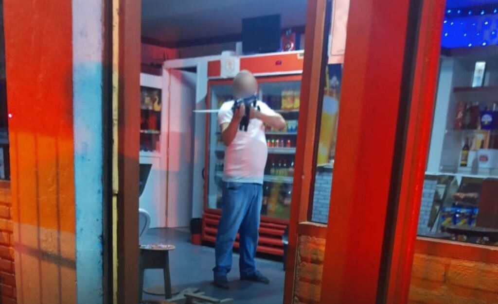 Homem de camisa branca e calça jeans segura arma apontada para policial. O suspeito está dentro de um bar e é possível ver geladeiras com produtos atrás dele.