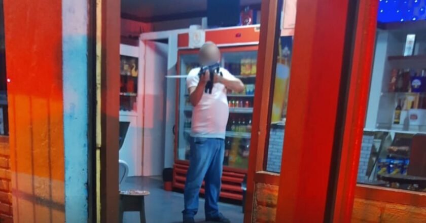 Homem de camisa branca e calça jeans segura arma apontada para policial. O suspeito está dentro de um bar e é possível ver geladeiras com produtos atrás dele.