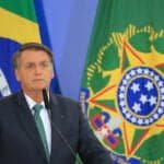Jair Bolsonaro, homem de pele branca, cabelos grisalhos na lateral da cabeça e cabelos penteados para o lado. Ao fundo bandeiras.