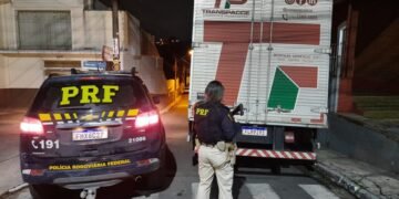 Viatura da Polícia Rodoviária Federal ao lado do caminhão. Agente da PRF, com arma longa, aparece olhando o caminhão, sobre a faixa de pedestres.