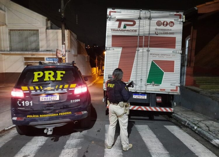 Viatura da Polícia Rodoviária Federal ao lado do caminhão. Agente da PRF, com arma longa, aparece olhando o caminhão, sobre a faixa de pedestres.