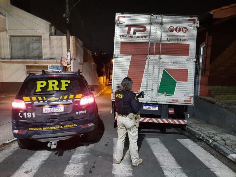Viatura da Polícia Rodoviária Federal ao lado do caminhão. Agente da PRF, com arma longa, aparece olhando o caminhão, sobre a faixa de pedestres. 