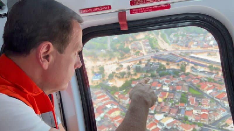João Doria, homem de pele clara e cabelos curtos, diante da janela do helicóptero aponta com o dedo região alagada de Franco da Rocha.