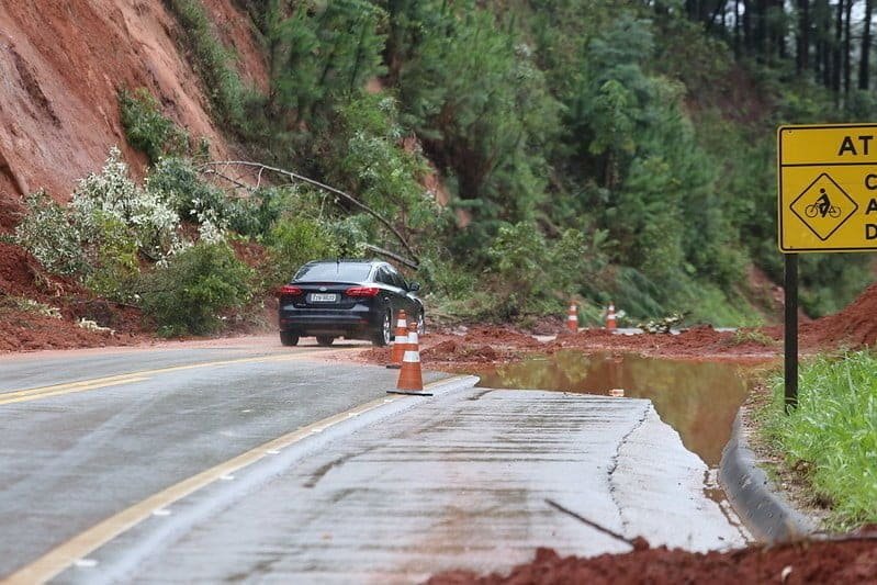 Imagem mostra carro passando por rodovia parcialmente encoberta por lama que deslizou da encosta. Cones sinalizam local tomado pela lama.