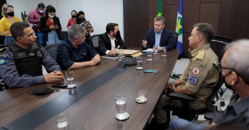 Governador Mauro Mendes, homem de pele branca e cabelos grisalhos, sentado à mesa, conversa com integrantes do governo e comando das polícias.
