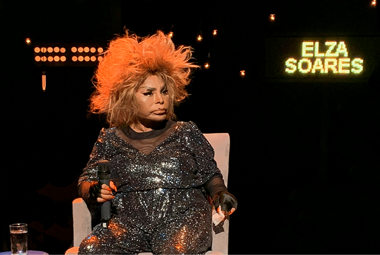 Elza Soares, mulher negra, usa macacão com brilhos, cabelo iluminados. Ao fundo, Elza Soares está escrito no cenário preto.