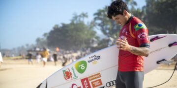 Gabriel Medina, de short e camiseta, segurando uma prancha de surfe, faz uma oração na areia da praia com a cabeça baixa e a mão esquerda levantada na altura do peito.