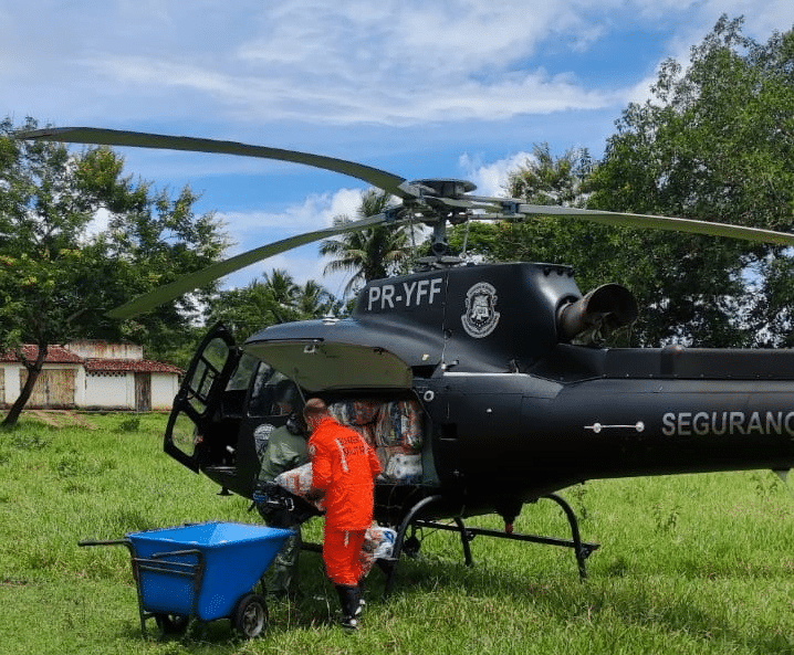 Helicóptero preto pousado na grama. Ao lado, com agente retira alimentos de dentro da aeronave e coloca e um carrinho de mão de cor azul.
