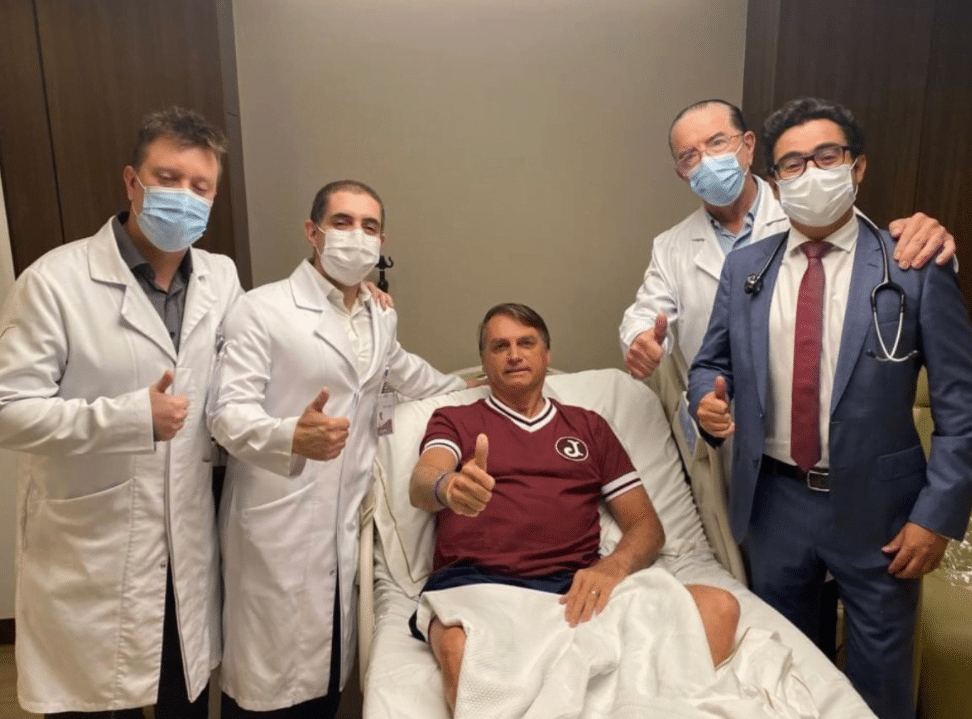 Jair Bolsonaro, homem de pele branca, sentado no leito de hospital fazendo gesto de positivo com a mão direita. Médicos ao lado, em pé, também fazem gesto de positivo.