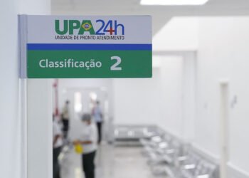 Foto mostra placa com logomarca da UPA 24 horas. Abaixo traz a indicação de "classificação 2". Ao fundo, desfocado, funcionários do hospital.