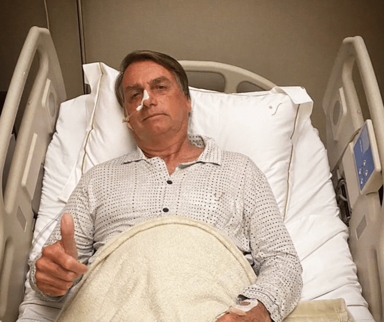 Jair bolsonaro, homem branco, deitado na cama de hospital, com sonda no nariz, faz gesto de positivo com a mão direita.