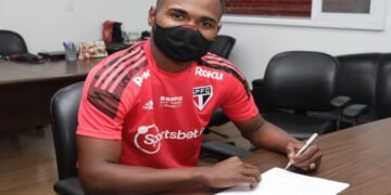 Jogador Nikão, usando uniforme do São Paulo Futebol Clube, segura a caneta diante de uma folha de papel colocada sobre a mesa. O jogador está com máscara.