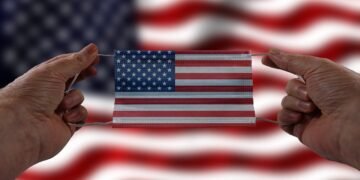 Bandeira dos Estados Unidos ao fundo, enquanto na frente duas mãos de uma pessoa branca seguram uma máscara em formato da bandeira americana.