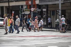 Imagem mostra pessoas caminhando na rua com máscara. É possível ver faixas de pedestre pintadas no asfalto.