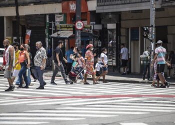 Imagem mostra pessoas caminhando na rua com máscara. É possível ver faixas de pedestre pintadas no asfalto.