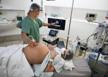 Gestante deitada, com a barriga exposta, enquanto médico observa no ultrassom os bebês em uma tela de computador.
