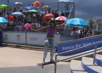 Rayssa Leal faz uma manobra aérea com skate, deslizando sobre barra de ferro . Ao fundo, público acompanha debaixo de guarda-sois coloridos.