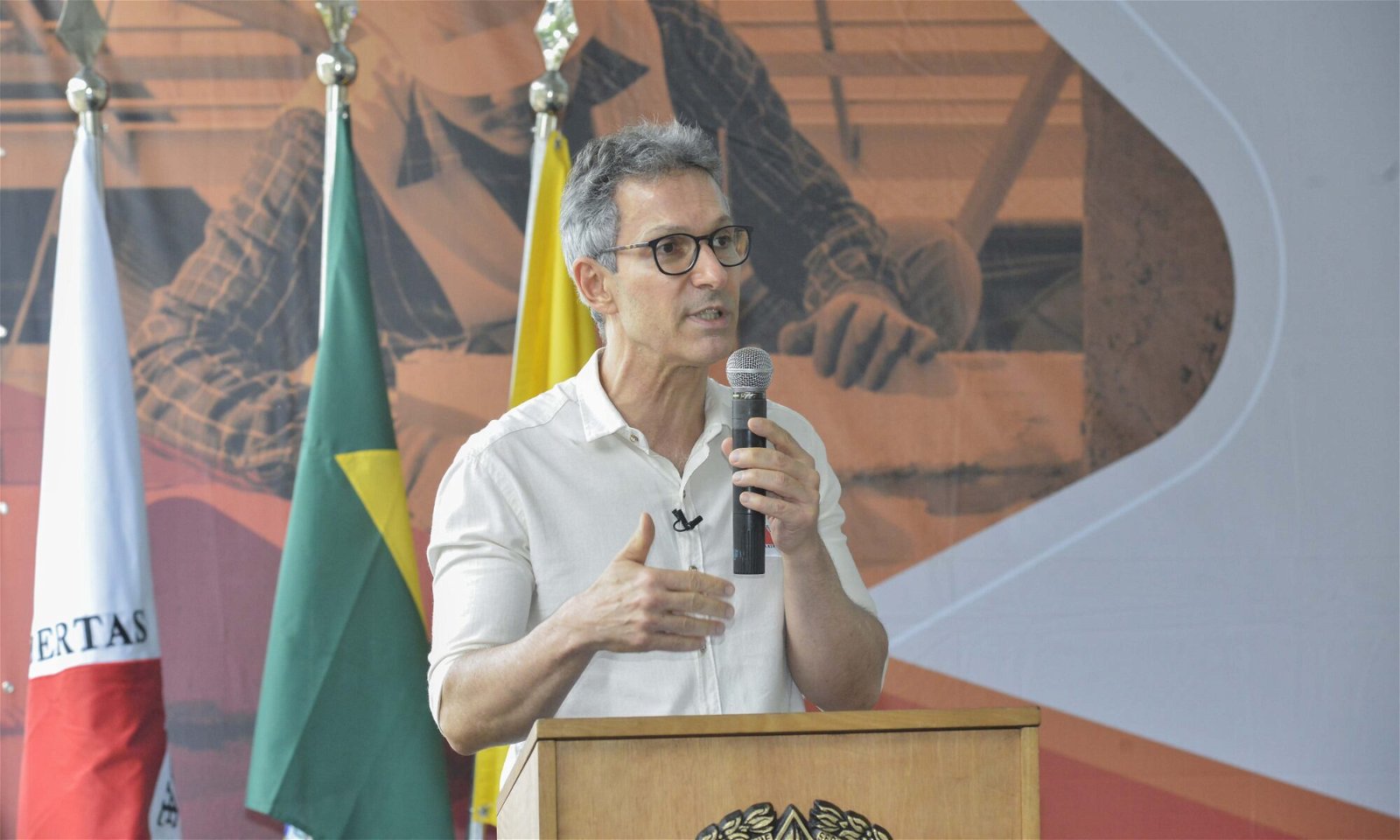 Romeu Zema, homem de pele branca, cabelo grisalho, segura microfone enquanto discursa. Ao fundo, bandeiras, inclusive do Brasil.