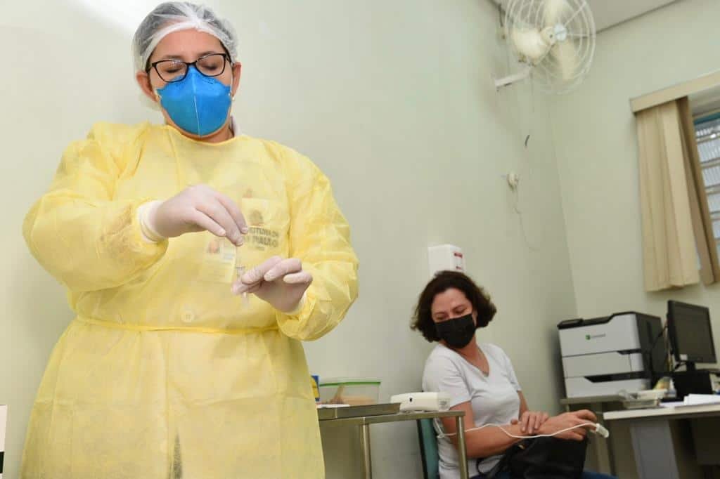 Agente de saúde com equipamentos de proteção no corpo, mãos, rosto e cabelo coleta amostra de exame enquanto paciente, uma mulher, usando máscara, aguarda sentada ao fundo.