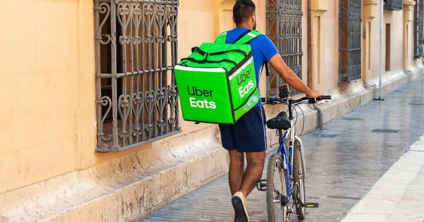 Com caixa de entrega nas costas, com logomarca da Uber Eats, entregador caminha sozinho por calçada enquanto empurra a bicicleta.